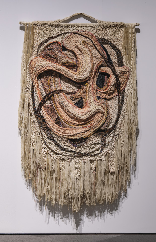 Photo: primarily tan wall hanging on a stick with a handle and a long fringe at the bottom. It is swirls in relief that have peach-orange and brown colouring. 