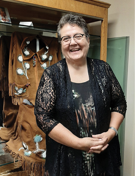 Colour photograph. An Indigenous woman is standing in front of a display cabinet holding a buckskin dress. She is smiling, with hands folded in front of her. She has greying curly short hair, wears glasses, and a black lace cardigan over a black patterned dress.