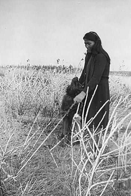 The image is a black-and-white photograph depicting a woman standing in a field of tall, dry grasses. The woman has long dark hair and is dressed in a long, dark coat or dress. She is interacting with a large wolf sitting in front of her, leaning into her legs and looking up at her. The background shows a vast field with scattered, tall plants, and a flat horizon. The image conveys a sense of calm and connection between the woman and the wolf within a natural setting.