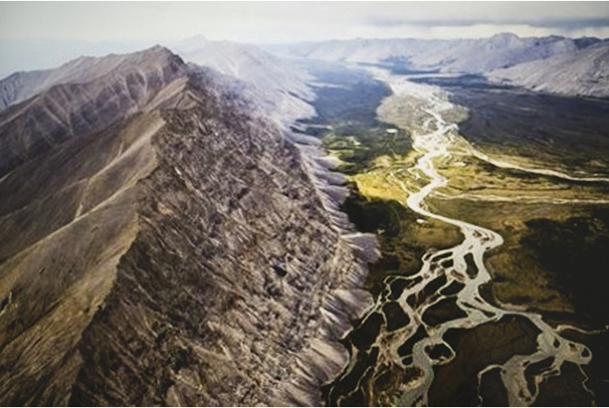 . An aerial view of the Taiga Cordillera ecozone of mountains spanning much of the northern Yukon. The rugged peaks and ridges stretch the length of the view and at the base of the peaks are rivers and creeks, flowing and taking direction from the flow from the top. Viewed from such a distance the rivers and creeks appear as small white ropes on the ground.