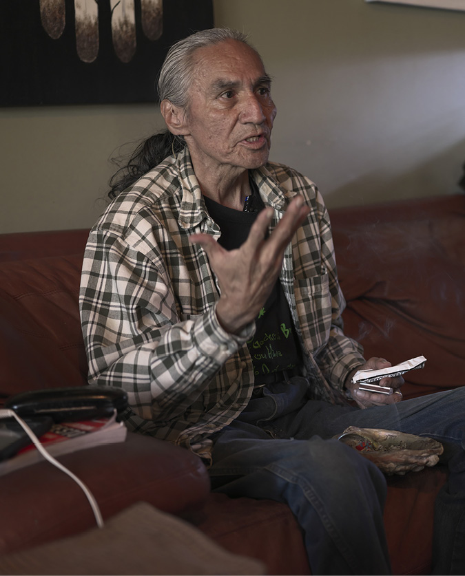 Dene Elder Patrick Deranger is seated, speaking with his right hand stretch out, and in his left hand is a ceremonial pouch of tobacco and matches.