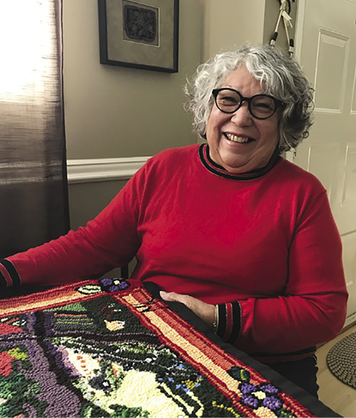 Smiling woman with curly grey hair and a red shirt sits in front of the rug from the previous image.