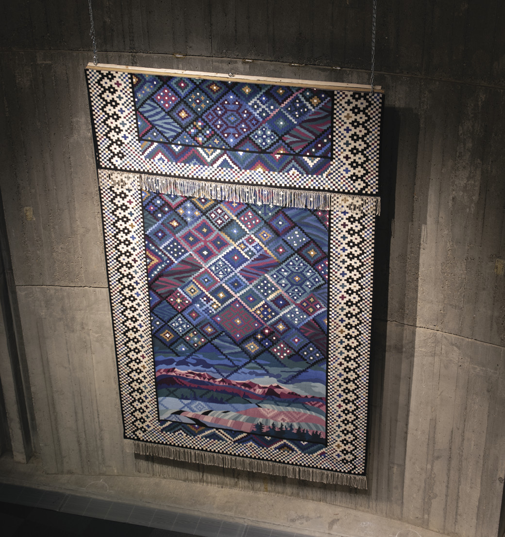 Photo: large blue hanging. It has a black and white patterned border and diamond in shade of blue, dark pink, green, and white in a pattern in the sky above pink-purple mountains and green, blue and pink hills. The piece is bisected across the top third with what could be the bottom of the piece repeated at the top.