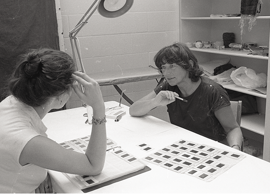 Black and white Photo: two women sit at a table talking with a binder and loose pages of slides on the table between them.