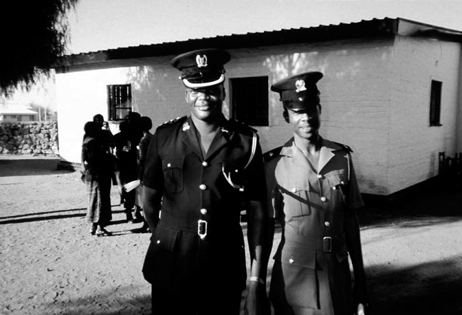A black and white photograph of Local police, Ramotswe, Botswana wearing police uniforms and a group of people chatting behind them.