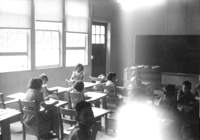 Black and white photograph: Twelve male and female children are seated at wooden desks in a classroom at the Mohawk Institute that was built from army huts.  One wall on the left side of the classroom consists of large windows and a door, and there is a desk and chalkboards at the front of the room. The photo is blurry and it is difficult to see faces, but the image provides a reasonable idea of what it was like inside a classroom at the school in the 1940s.
