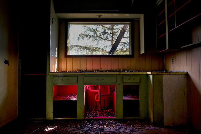 Sunlight beams into a kitchen window of a long-abandoned house revealing a kitchen counter frame with wooden cupboard doors removed. Front and centre, in the cupboard below the window, are water pipes under the space where a sink and taps are removed. The streaming sunlight reveals dust and scattered leaves on the surfaces and floor. The shelves on the walls to the right are stripped bare. This image was captured in an abandoned 1950s home in Uranium City, Saskatchewan.