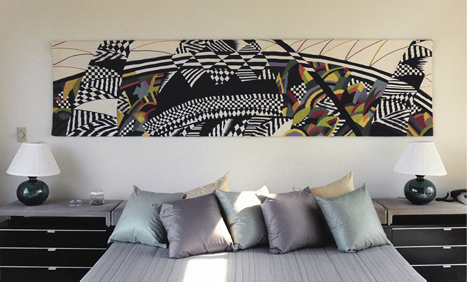 Photo: a bed piled with green and grey pillows between two black nightstands with green lamps. Above the bed hangs a long piece of art. It is mostly black and white with waves of green-yellow-red-orange.