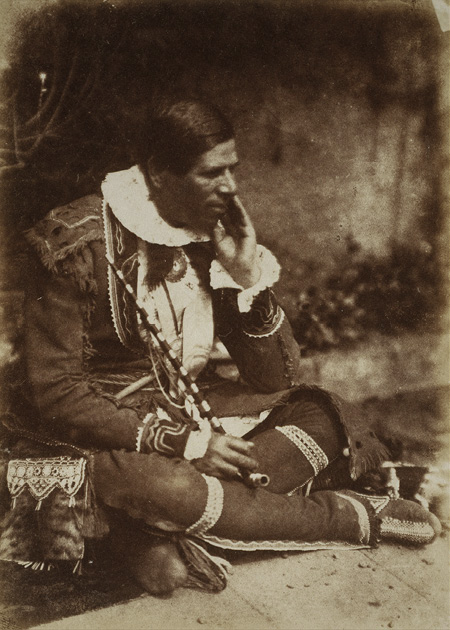 Black and white historical photograph. A man in Indigenous regalia is sitting cross-legged. He is wearing an elaborately decorated jacket, leggings, and moccasins. He is holding a long-stemmed pipe in his right hand, and touching his face with his left. He has a decorated bag hanging to his side.