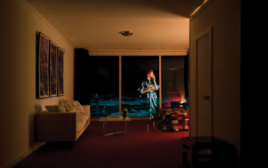 Colour photograph: A diorama of a bedroom in a suburban home, with a projected image of man pointing to something on the television.