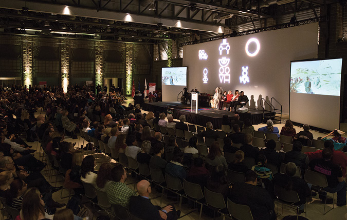Colour Photograph: Large audience participating in Indigenous Strategy launch event, with videos playing on left and right side of the stage and cultural symbols in the centre.