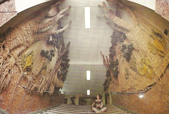 Photo: a woman sits on stairs looking up at walls lined with 3D trees and foliage that curve over her. 
