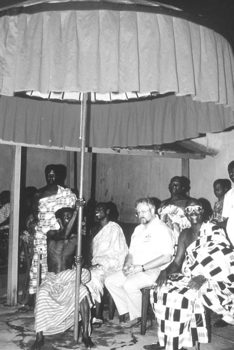 A black and white photograph with a person holding a giant umbrella and some people are sitting and some are standing in the front of the umbrella.