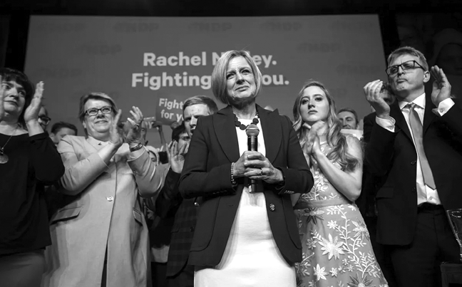 Rachel Notley stands indoors in from of a sign partly obscured by her head, but which reads “Rachel Notley. Fighting for you.” She is holding a microphone with both hands. She is surrounded by people clapping.