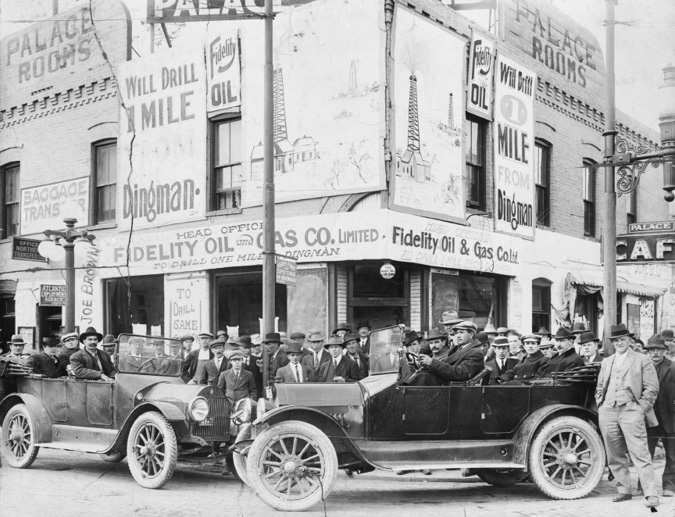 Figure 5-10 “Men in car and on sidewalk waiting to invest in oil stock.” Black and white photograph featuring a large crowd of spectators gathered outside the Palace Rooms Hotel on Atlantic Avenue. One newspaper noted that the boom altered the city’s skyline as businesses quickly fixed large hand-printed signs to the exterior of buildings advertising individual companies, and this can be seen in this image as the Palace also served as the headquarters for Fidelity Oil and Gas Company. Two early-model, open top automobiles are parked in front of the hotel. In the car on the left, driller Joe Brown is identified by a written note on the photograph.