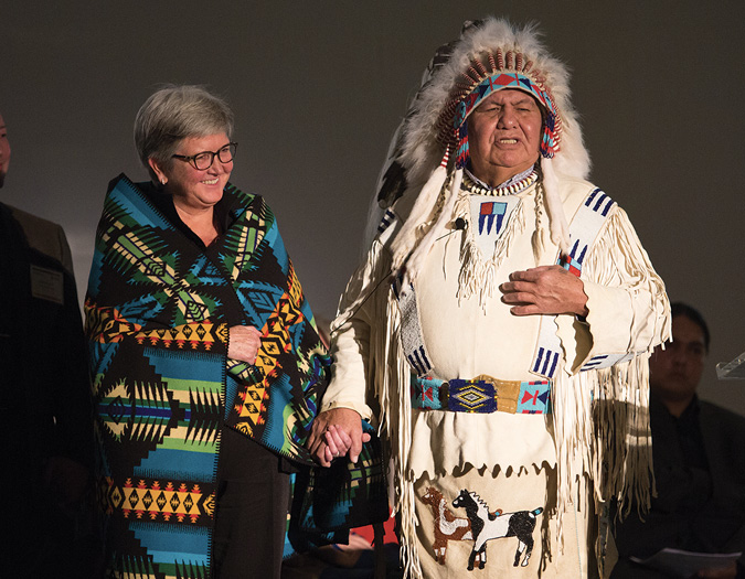 Colour Photograph: Dr. Reg Crowshoe in traditional regalia, gifting Blackfoot name to Dr. Dru Marshall, wearing a traditional Pendleton blanket.