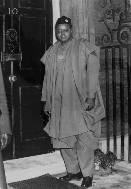 A black and white photograph of Dr. M. I. Okpara wearing a huge jacket and a cap while standing in front of a door.