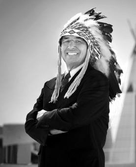 Black and white photograph: A man dressed formally in a suit and tie, wearing a traditional feathered headdress, stands with arms crossed, smiling broadly. A tipi is in the background to the right.