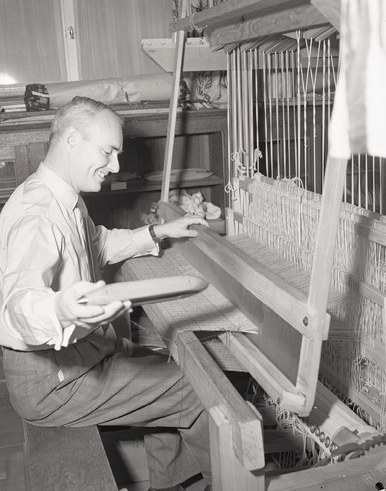 Black and white photograph of a man sitting at a look, holding out a tool toward the picture taker.