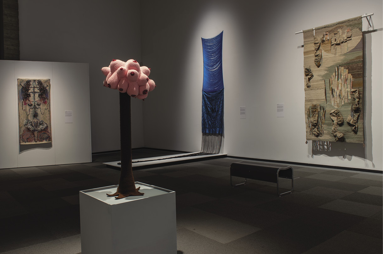 Four pieces of art in a gallery. Far left: a weaving of a distorted face. Left-middle: a profusion of pink blobs with red nipples at the top of a brown woven tree trunk. Right middle: an extra tall blue hanging with horizontal string in the top half, solid blue in the bottom half, and a dark blue fringes at the bottom. Right: a tan, sage-green, and brown piece with 3d elements. 