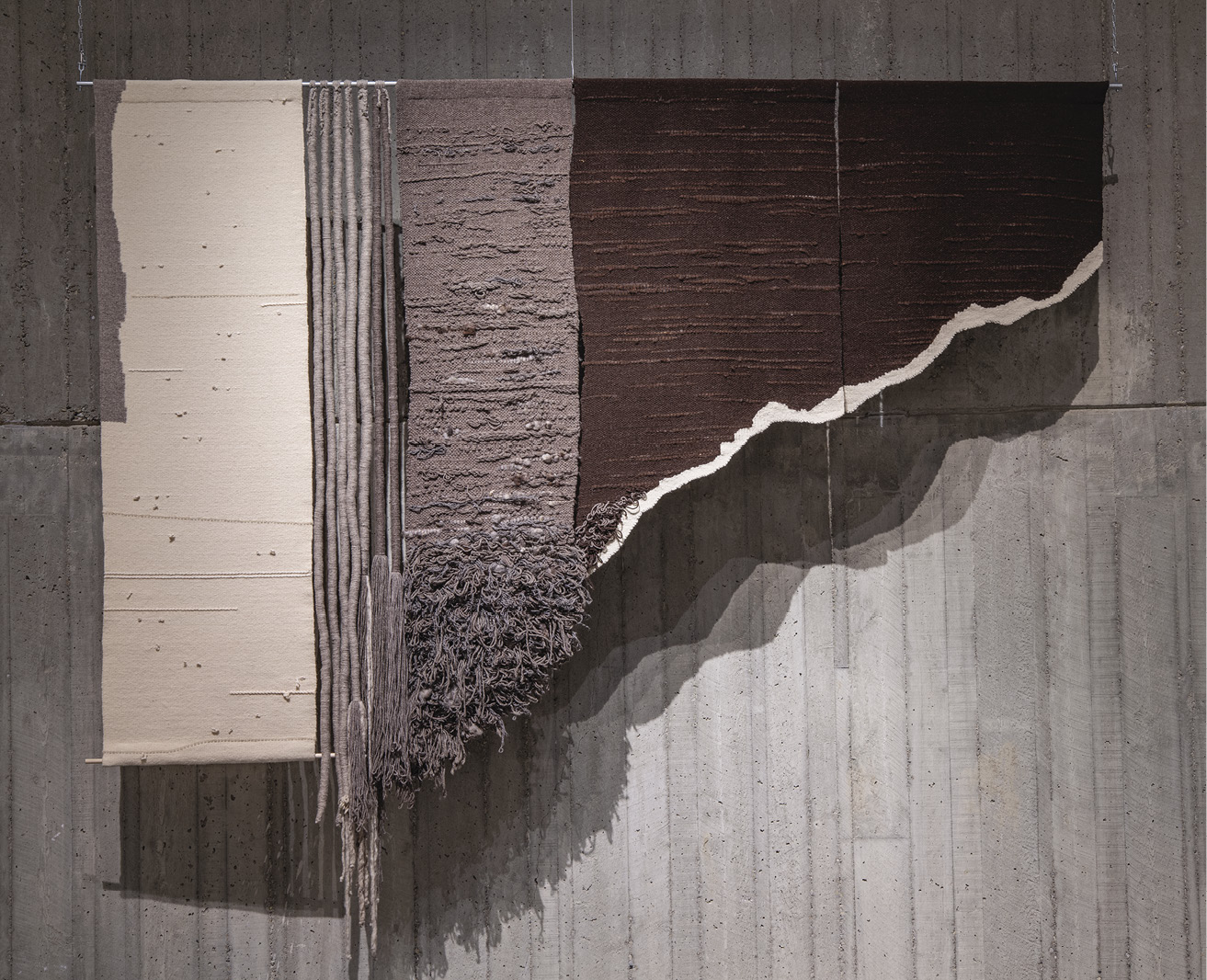 Photo: large angular hanging piece made up of several panels — along tan panel, six light grey ropes, a wide grey 3d panel with a messy fringe, and two darn brown panels with a torn off, angled edge.