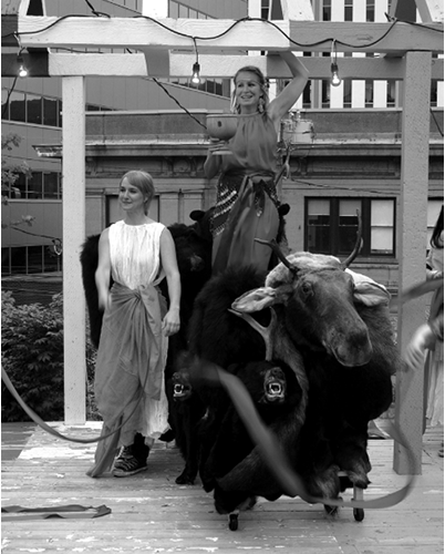 Black and white photograph, two women in togas stand performing beneath a pergola, the woman on the right is holding a large goblet and stands atop a cart made of a taxidermized moose head and two taxidermized bear heads, hands reach in from outside the photograph waving ribbon streamers.