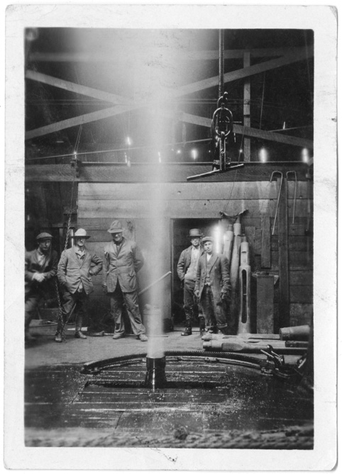 Figure 1-7 “Group of men standing at well-head of Dingman #1” Black and white picture of five men inside the drill shack of the Dingman #1 well watching the well vent petroleum condensate. None of the men are wearing any protective clothing or safety gear.