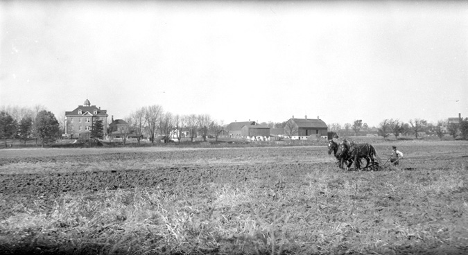 Black and white photograph: Distant view of the outside of the third Mohawk Institute building surrounded by large farmlands.  The school is bordered by trees and several outbuildings and barns.  In the foreground, there are two horses pulling a plow controlled by a male figure.