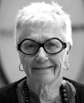 Black and white photograph: A portrait of an older woman with short white hair, wearing thick round glasses. She is smiling gently and looking directly at the camera.