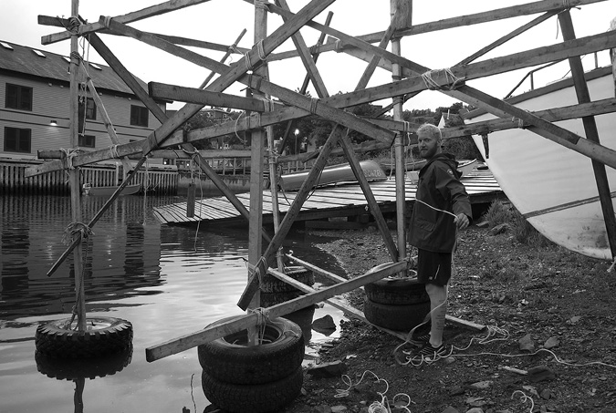 Black and white photograph, a man in shorts and windbreaker roped together a wooden scaffold, sitting on tyres at the edge of the water, a boat ramp with canoes and a large wooden building sit in the background.
