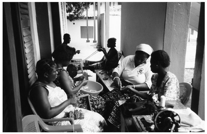 A black and white photograph of about seven women from Manya Krobo Queenmothers Association making bead necklaces and bracelets, sitting in a room.