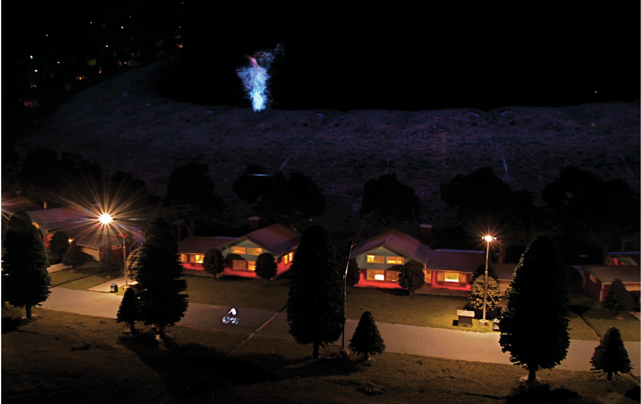 Colour photograph: A diorama of a suburban street with houses at night, with a projected figure of a cyclist riding on the street in the foreground.