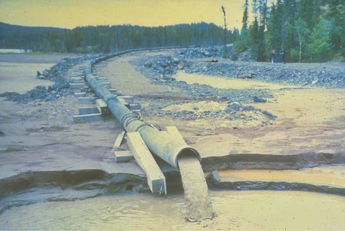 An long steel pipe propped up through a distance through a man-made land path through forested areas and waterways to Fookes Lake provides an outlet for waste water.