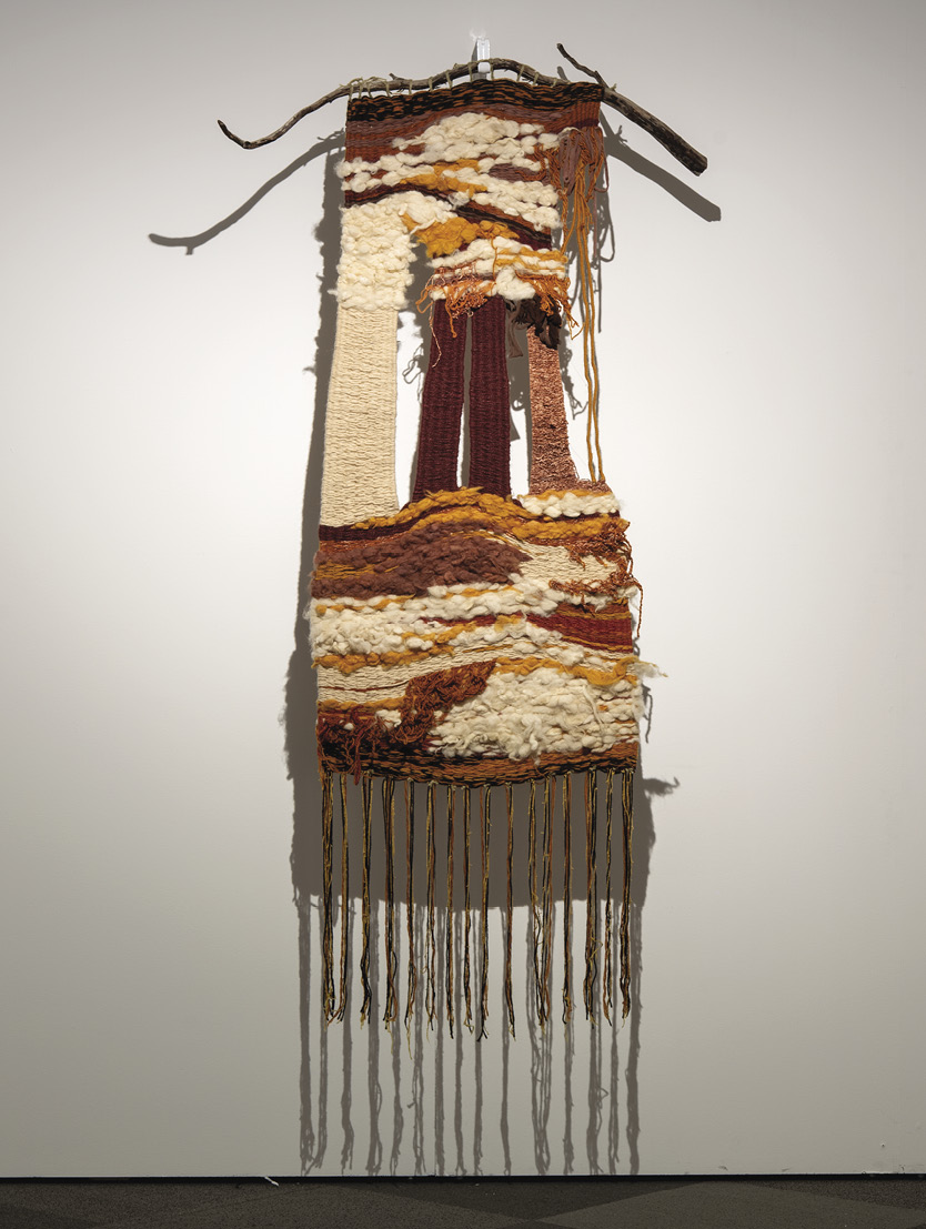 Photo : un textile marron, blanc cassé et orange suspendu à un bâton incurvé sur un mur blanc. Des ficelles artisanales partent du bas.