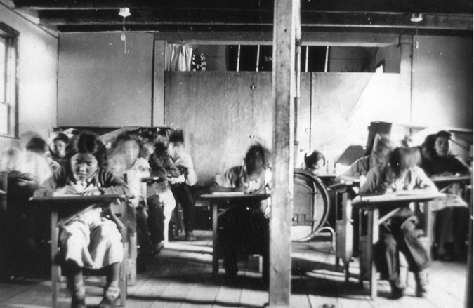 The black and white image depicts a classroom setting with several children seated at individual wooden desks. The room is dimly lit, with natural light streaming through a window on the left. The children appear focused, engaged in writing tasks. The walls and ceiling are plain, with exposed wooden beams. A large wooden post is centrally positioned within the room, dividing the space. The floor is unadorned and wooden, adding to the simple, utilitarian decor. The overall tone of the image is slightly blurred, suggesting motion from the seated children.