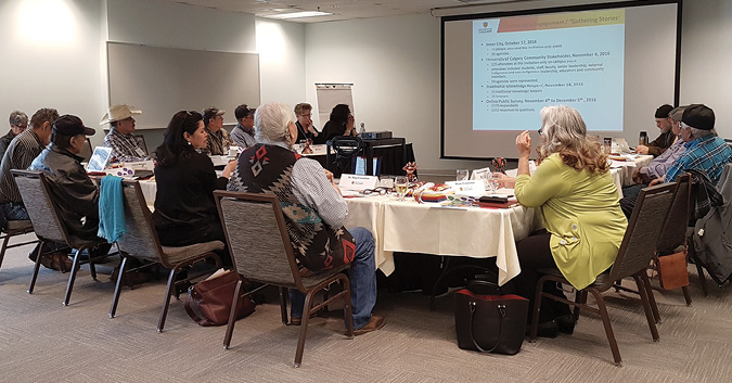 Colour Photograph: Traditional Knowledge Keepers sitting at a table in a room with a projector.