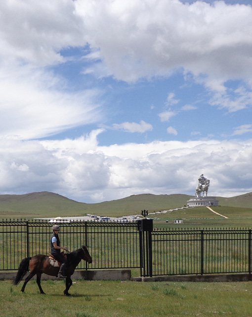 Mounted rider on takhi walking past fence and monument