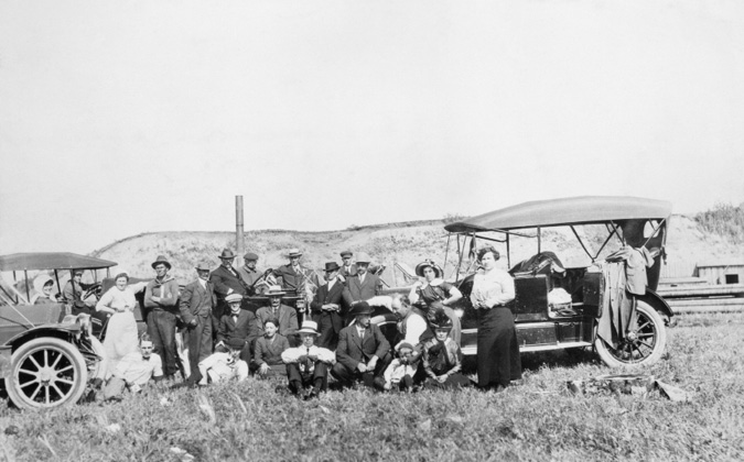Figure 6-2 “Picnic group at site of Dingman #1 well.” Black and white photograph of a picnic at the Dingman n#1 well in Turner Valley, where a glimpse of one of the buildings is visible behind the automobile on the right. The image is dominated  by a group of men, women, and children dressed casually but properly, in long dresses, hats, suits, and ties. The picture also features two early-model open-top automobiles with one draped in fabric or clothing, possibly for shade or storage.