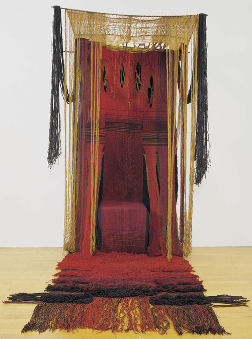 Photo: a piece of art that almost looks like a booth. There is a red backing with black and gold feathers hanging down, a roof constructed of threads strung across and down the sides, and then a red and black rug with a long fringe laying in front of the structure.