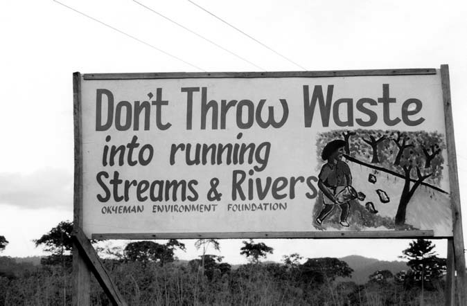 A black and white photograph of a slogan on the billboard and the slogan being 'Don’t Throw Waste into running Streams and Rivers' with a small picture of a woman throwing waste into a river.