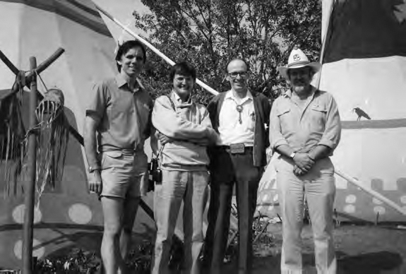 A photograph of four people with a tent and trees in the background.