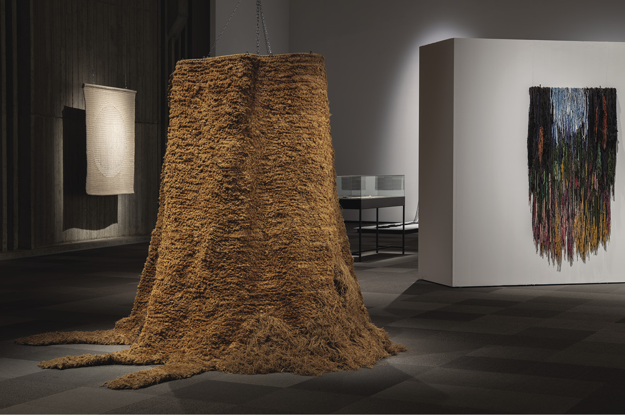 Large shaggy brown artwork in the shape of a 10-foot-tall tree stump flanked by two spotlighted pieces — once cream coloured and portrait-orientation rectangle, the other a blue-black-green-orange hanging — in a gallery setting.