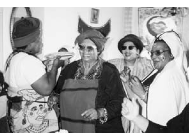 Three smiling women are dressed in traditional attire inside a restaurant. Another woman is carrying a plate. On the wall behind them, a mirror and a couple of paintings are hung.