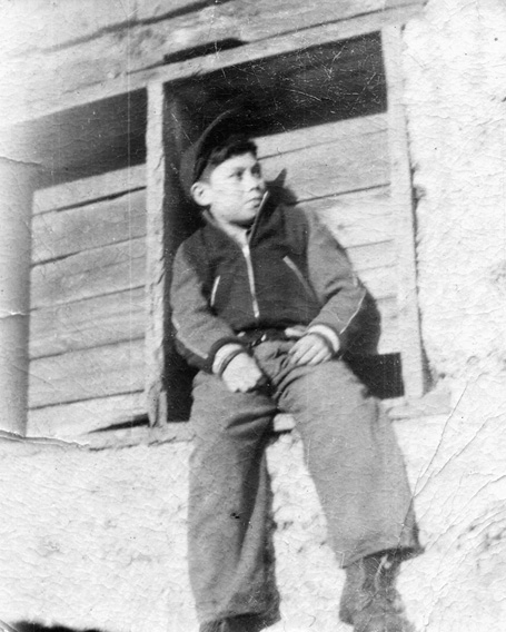 Black and white photograph: Young Indigenous male in casual clothing and a hat sitting in what appears to be a framed opening in a wall.  He is looking off to the right.