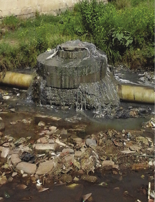 Colour photograph: Photo of a sewer main pipe outdoors, above-ground, with a clogged valve or grate, surrounded by garbage and dirty water.