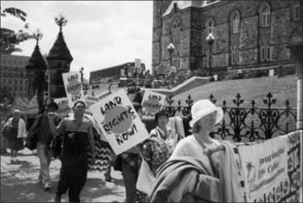 Men and women are standing outside a grand castle and carrying signs that read, Land Rights Now!