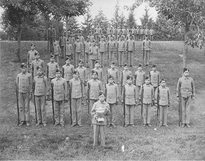 Black and white photograph: Five rows which add up to approximately fifty Indigenous male cadet students formally posed outdoors on a grassy hill. There is one boy in front holding a trophy.  All of the students are wearing cadet uniforms and caps and holding firearms at their sides.