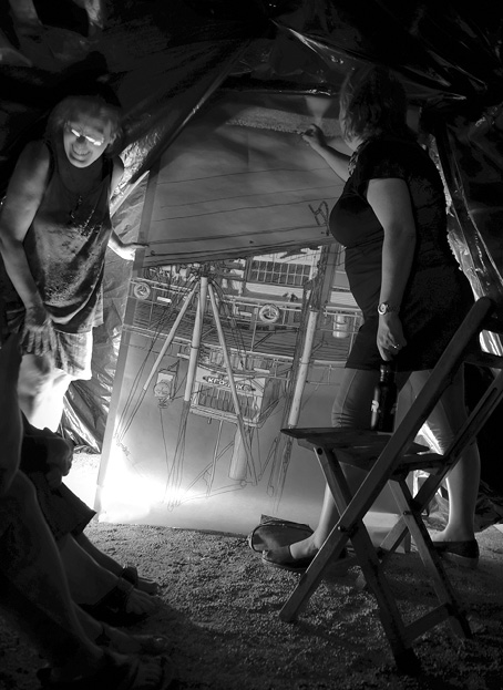Black and white photograph, two women inside a dark tent hold a large canvass decorated with an upside down image of a paddle steamer, the SS Klondike; a fold-up chair and several feet are visible in the foreground.