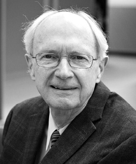 Black and white photograph: A portrait of an older man wearing glasses and a suit and tie. He is smiling gently and looking directly at the camera.