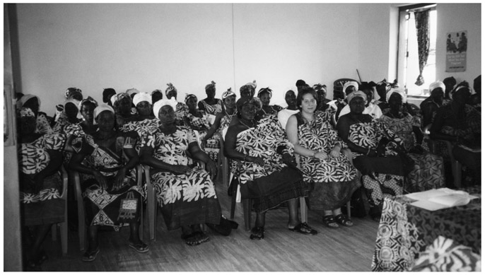 A black and white photograph of Kimberley Schoon, now Manye Pi Yo Gu, sitting with Manye Nartekie, Paramount Queenmother and other Queenmothers after installation as Honorary Queenmother. There are around four rows of women dressed similarly, sitting in a room in front of the dais.